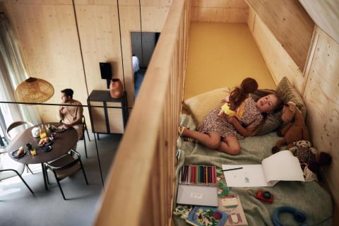 Un hombre desayuna en una mesa mientras una niña juega con una muñeca en el altillo de una tiny house en Holanda.