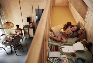 Un hombre desayuna en una mesa mientras una niña juega con una muñeca en el altillo de una tiny house en Holanda.