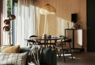 Acogedor interior de casa pequeña con paredes de madera, mesa de comedor, sofá y luz natural de grandes ventanas.
