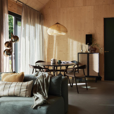 Cozy tiny house interior featuring wood-paneled walls, dining set, sofa, and soft daylight through large windows.