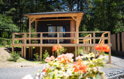 Edelweiss-houten lodge met groot terras in een bosrijk gebied, bloemen op de voorgrond, zonnig weer.