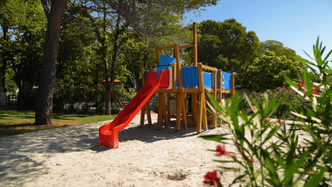 Parco giochi con scivolo rosso e sabbia, circondato da alberi, al Camping Zelena Laguna, Istria, Croazia.
