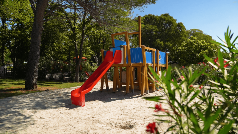Spielplatz mit Rutsche im Sand, umgeben von Bäumen, aufgenommen im Camping Zelena Laguna in Istrien, Kroatien.