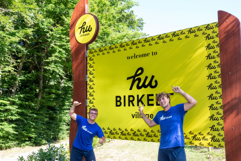 Deux personnes en t-shirts bleus 'hu team' posent devant le panneau d'accueil du hu Birkelt Village au Luxembourg.