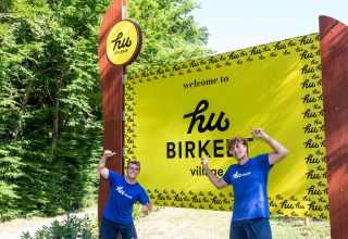 To personer i blå 'hu team' trøjer foran velkomstskiltet til hu Birkelt Village feriepark i Mersch, Luxembourg.