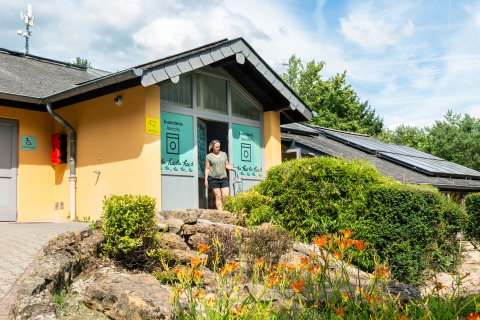 Kvinde forlader vaskerum ved hu Birkelt Village feriepark i Mersch, Luxembourg, omgivet af blomster og grønt.