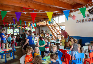 Children and adults join craft activities under colorful bunting in a busy hall at hu Birkelt Village, Luxembourg.