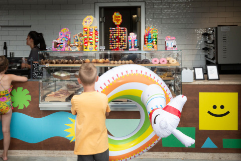 Children in swimwear choose pastries and candies at a colorful cafe in hu Birkelt Village, Luxembourg.