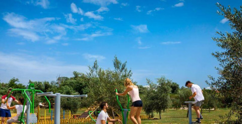 People exercising outdoors on fitness equipment at Camping Aminess Maravea, Istria, Croatia, under blue sky.