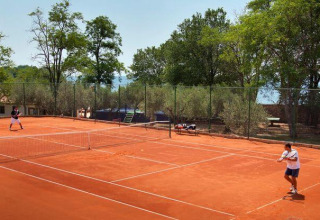 To personer spiller tennis på en rød grusbane omgivet af træer i Camping Aminess Maravea, Istrien, Kroatien.