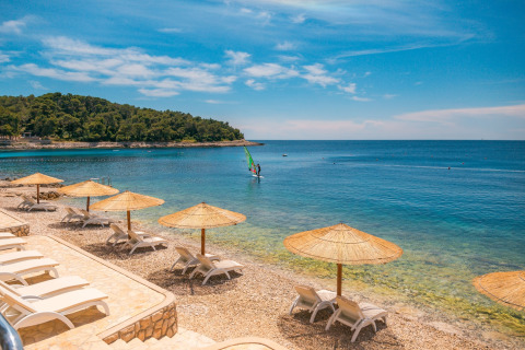 Ligstoelen en rieten parasols op het strand bij Camping Cikat, Primorje-Gorski Kotar, Kroatië, onder blauwe lucht.