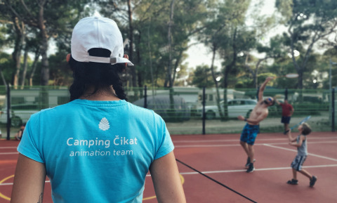 A person in a Camping Čikat shirt watches people playing badminton on an outdoor court among trees.