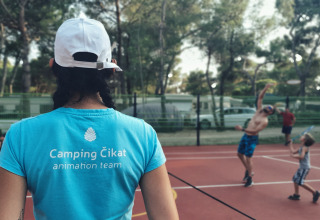 En person med Camping Čikat t-shirt ser på badmintonspillere på en udendørs bane blandt træer.