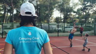 Persona con camiseta de Camping Čikat observa a gente jugando bádminton en una pista al aire libre.