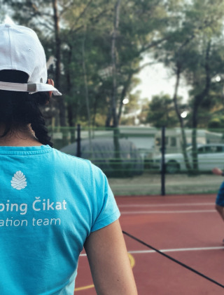 Persona con camiseta de Camping Čikat observa a gente jugando bádminton en una pista al aire libre.