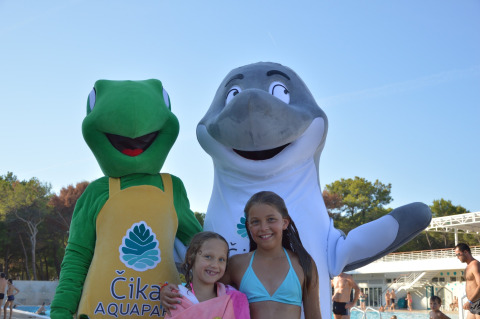 Two children pose with a frog and dolphin mascot by the pool at Camping Čikat holiday park in Croatia.