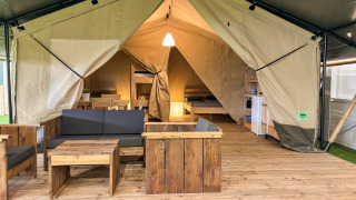Interno tenda safari con arredi in legno, area salotto, letto e angolo cottura su pavimento in legno.