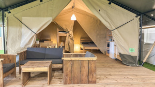 Interno tenda safari con arredi in legno, area salotto, letto e angolo cottura su pavimento in legno.