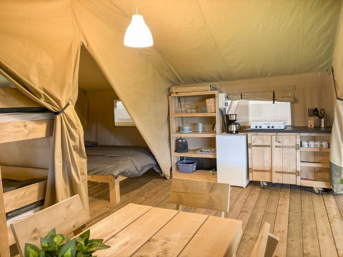 Interior de tienda safari con litera, cama doble, mesa de comedor y cocina totalmente equipada en suelo de madera.
