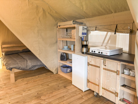Interior de una tienda safari con suelo de madera, cama y pequeña cocina equipada con nevera y placa.