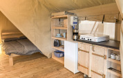 Interior de una tienda safari con suelo de madera, cama y pequeña cocina equipada con nevera y placa.