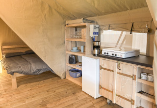 Interior de una tienda safari con suelo de madera, cama y pequeña cocina equipada con nevera y placa.