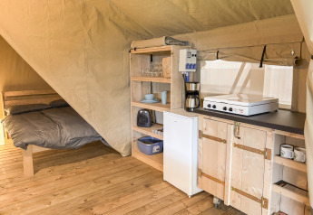 Interno di una tenda safari con pavimento in legno, letto e piccola cucina attrezzata con frigo.