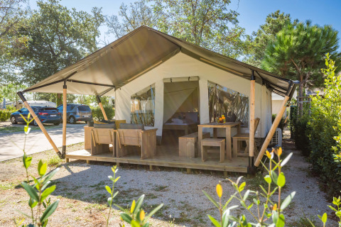 Tienda Safari Comfort con aire acondicionado, terraza amueblada en camping soleado, coches al fondo.