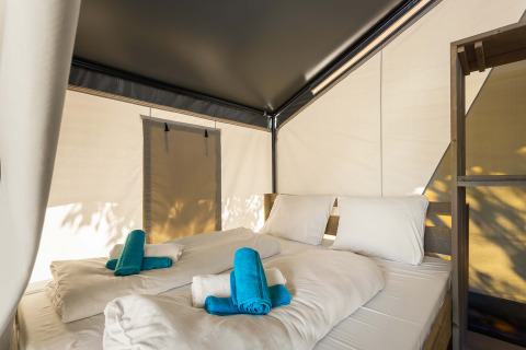 Interno di una tenda safari con letto matrimoniale, biancheria bianca e asciugamani turchesi.