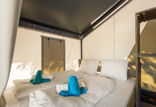 Interior de una tienda safari con cama doble, ropa de cama blanca y toallas turquesas encima.