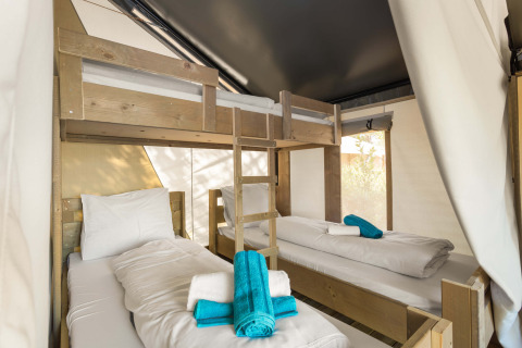 Vista interna di una tenda safari con un letto a castello e due singoli, biancheria bianca e asciugamani blu.