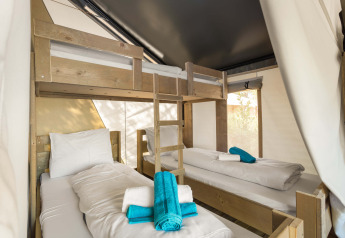 Interieur van safaritent met stapelbed en twee eenpersoonsbedden, opgemaakt met witte lakens en blauwe handdoeken.