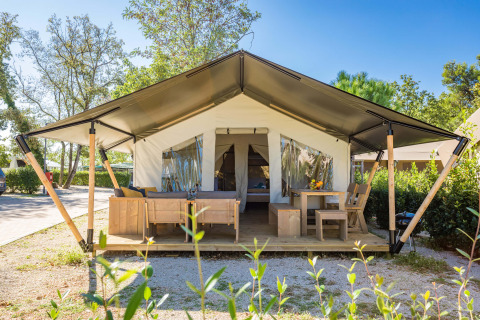 Vista exterior de Safari Tent Comfort con muebles de madera en la terraza y entrada abierta rodeada de plantas.