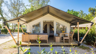 Safari Tent Comfort von außen mit Holzmöbeln auf der Veranda und offenem Zelteingang, umgeben von Bäumen.