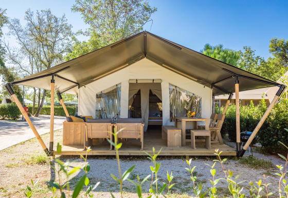 Vista exterior de Safari Tent Comfort con muebles de madera en la terraza y entrada abierta rodeada de plantas.