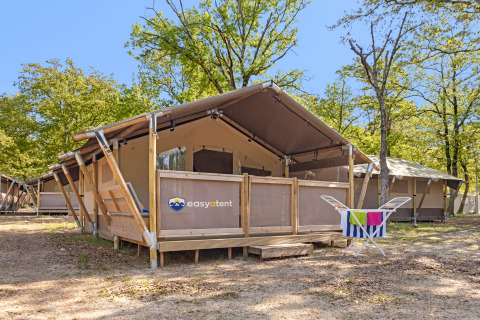 Tente lodge safari sur un camping boisé avec véranda et étendoir à linge portant des serviettes colorées.