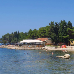 Camping Stella Maris en Istria, Croacia, con bañistas, paseo marítimo y árboles bajo un cielo azul despejado.