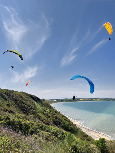 Vier paragliders zweven boven groene heuvels en strand bij Toftum Bjerge Camping in Denemarken.