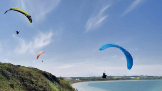Fire paraglidere svæver over de grønne bakker og stranden ved Toftum Bjerge Camping i Danmark.