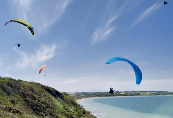 Four paragliders soar over green hills and coastline at Toftum Bjerge Camping in Central Denmark.