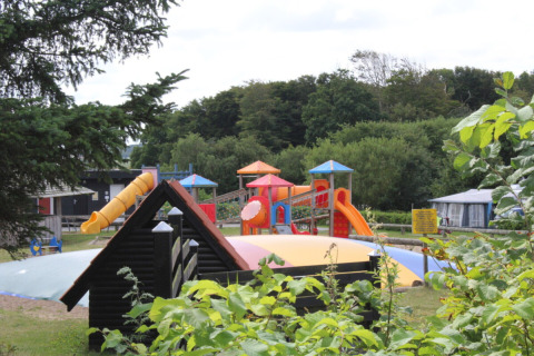 Parco giochi con scivoli e tappeto elastico a Toftum Bjerge Camping, circondato da alberi e cespugli.
