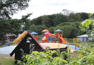 Playground with slides and a jumping pillow at Toftum Bjerge Camping, surrounded by trees and bushes.