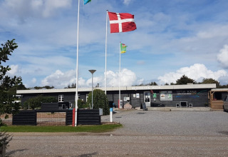 Toftum Bjerge Camping i Danmark med dansk flag foran informationsbygning og parkeringsplads i solskin.