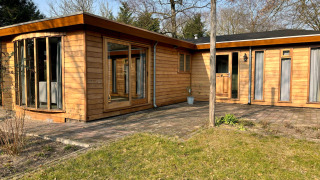 Vue extérieure de Cozy Lodge en bois, grandes baies vitrées et terrasse ensoleillée dans un cadre paisible.