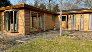 Udendørsbillede af Cozy Lodge med træfacade, store vinduer og solrig have i fredelige omgivelser.