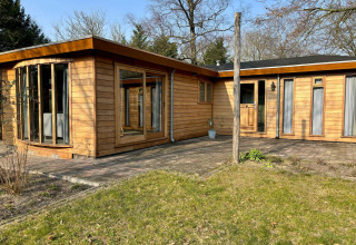 Udendørsbillede af Cozy Lodge med træfacade, store vinduer og solrig have i fredelige omgivelser.