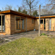 Udendørsbillede af Cozy Lodge med træfacade, store vinduer og solrig have i fredelige omgivelser.