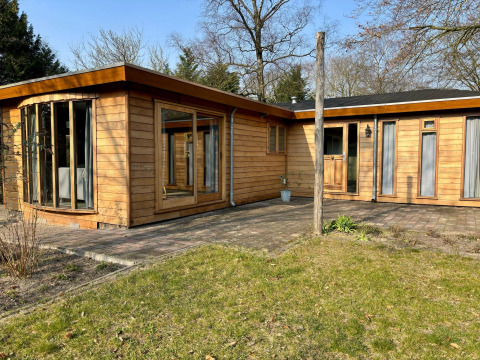 Buitenaanzicht van Cozy Lodge met houten bekleding, grote ramen en zonnig terras in een rustige sfeer.