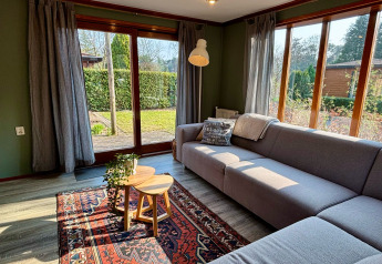 Soggiorno accogliente di lodge con ampie finestre, divano grigio e vista giardino a Het Verscholen Dorp.