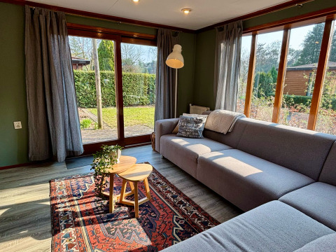 Soggiorno accogliente di lodge con ampie finestre, divano grigio e vista giardino a Het Verscholen Dorp.